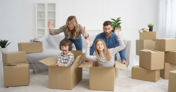 젊은 백인 부모들은 새 집으로 이사하는 동안 아이들과 집에서 즐겁게 지낸다. 상자에 앉아서 엄마 아빠랑 아파트에서 노는 동안웃고 있는 어린 아이들, 소년 소녀들 — 비디오