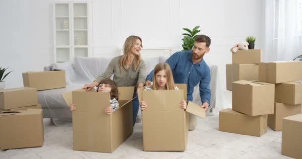 Padres caucásicos felices divirtiéndose en la sala de estar con los niños mientras se mudan a una casa nueva. Lindos niños pequeños, niño y niña sentados en cajas de cartón y jugando con la madre y el padre en un apartamento nuevo. — Vídeos de Stock