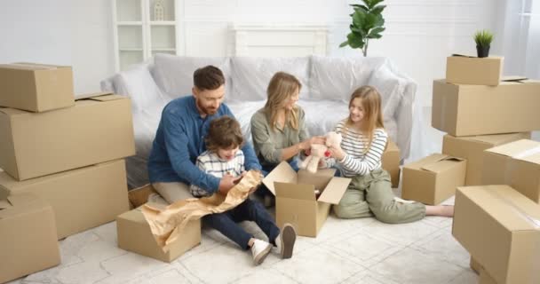 Veselí bělošští rodiče sedí na podlaze v obývacím pokoji s malými roztomilými dětmi a vybalují kartónové krabice. Rodina s dětmi stěhování v novém domě po opravě Chlapec a dívka s matkou ad otec — Stock video