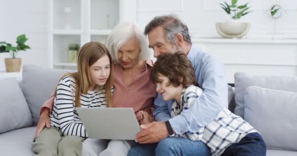 Kaukasiska äldre morföräldrar med barnbarn sitter på soffan hemma och använder bärbar dator. Mormor, farfar, barnbarn och barnbarn spenderar tid tillsammans på internet. Inomhus. — Stockvideo