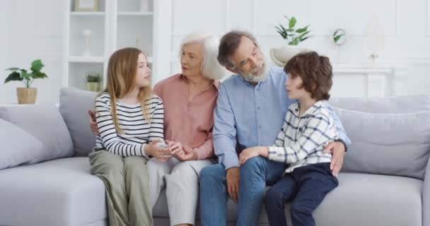 Kavkazští staří prarodiče s vnoučaty sedí na gauči v útulném obývacím pokoji. Babička, dědeček, vnuk a vnučka spolu tráví čas doma. Dovnitř. Rodinné generace. — Stock video