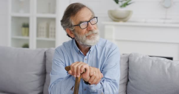 リビングルームの棒でソファに座っている間、白人の老練な賢いハンサムな男の肖像画は、側を見て、眼鏡で派手そうな目で考えています。杖を持つ重大な祖父。休みの日 — ストック動画