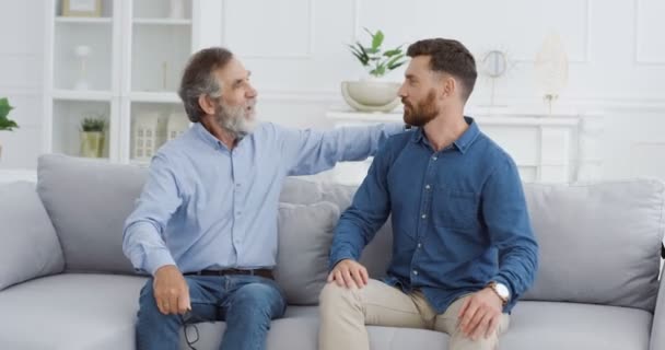 Hombres caucásicos generaciones de la familia. Feliz padre mayor alegre abrazando a su hijo guapo adulto en el sofá en casa. Dos hombres de diferentes edades riendo y pasando tiempo juntos en la sala de estar. — Vídeos de Stock