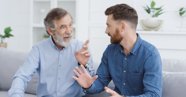 Famille masculine blanche générations. Heureux père sage aîné avec les cheveux gris dire quelque chose au fils adulte et expliquer sur le canapé dans le salon confortable. Vieil homme enseignant et prêchant jeune homme à l'intérieur. — Video