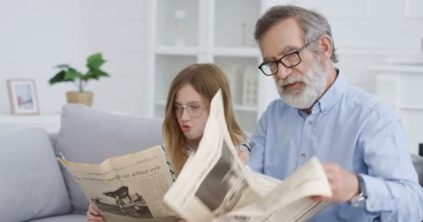 Caucasian old wise grandfather in glasses with cute small teen daughter sitting on couch in cozy living room and reading newspaper. Senior gray-haired man grandpa read papers with nice little girl. — Stock Video