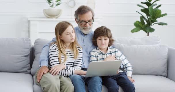 Bělošský děda sedí na gauči se dvěma malými dětmi a povídá si. Dědeček objímá malé děti na gauči a používá společně notebook. Doma. Starší muž v brýlích s vnoučaty. — Stock video