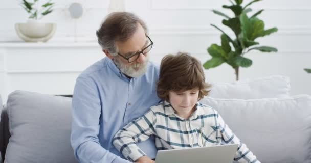 Biały dziadek siedzi na kanapie w salonie i uczy małego uroczego wnuka za pomocą laptopa. Mały chłopiec pisze na komputerze i gra w grę. W środku. Dziecko spędza dzień z dziadkiem, wygrywając. — Wideo stockowe