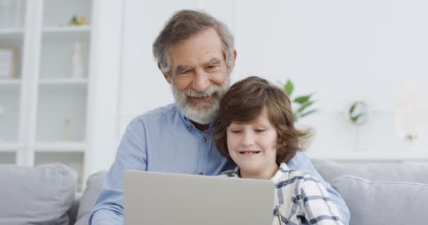 Běloch dědeček sedí na gauči v obývacím pokoji a učí malého roztomilého vnuka používat notebook. Malý chlapec píše na počítači a hraje hru. Dovnitř. Den strávený dětmi s dědou, smějící se. — Stock video