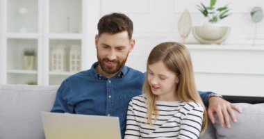 Yakışıklı beyaz baba oturma odasında kanepede oturuyor ve küçük şirin kıza dizüstü bilgisayar kullanmayı öğretiyor. Küçük kız babasıyla bilgisayarda oynuyor ve izliyor. Video. Çocuk babayla bir gün geçiriyor.