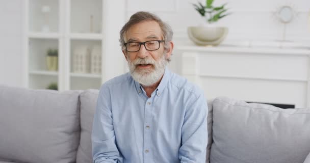 Retrato del guapo viejo bloguero caucásico en gafas sentado en el sofá en la sala de estar y hablando con la cámara. Vlogger masculino diciendo algo y explicando. Video blogging. Vlog desde casa Videochat — Vídeo de stock