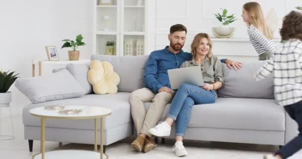 Two cheerful Caucasian kids running in circles and rounds in living room. Happy cute small sister and brother playing around sofa. Smiled mother and father sitting on couch with laptop computer. — Stock Video