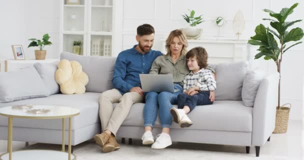 Jonge blanke ouders die thuis op de bank zitten en kleine schattige zoon les geven met een laptop. Kleine jongen met moeder en vader kijken iets op de computer in de woonkamer. — Stockvideo