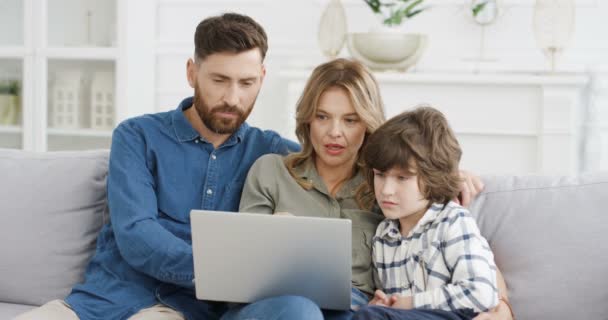 Küçük çocuklu beyaz ebeveynler evdeki kanepede oturup dizüstü bilgisayarı tıkırdatıp oynatıyorlar. Anne ve baba küçük oğluna oyun oynamayı ve bilgisayar kullanmayı öğretiyor ve birlikte video ailesi gösteriyorlar. — Stok video