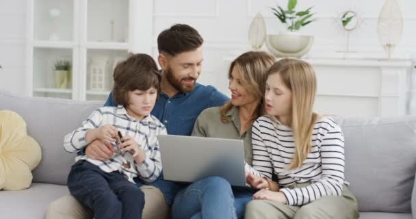 Szczęśliwe małe nastolatki siedzące w uściskach z rodzicami na kanapie i zakupy online z karty kredytowej na laptopie. Matka ojciec z małymi dziećmi, córka i syn kupują w internecie i płacą. — Wideo stockowe