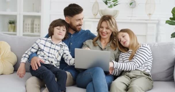 Niños adolescentes pequeños y felices sentados en abrazos con los padres en el sofá y viendo videos en la computadora portátil. Madre padre con niños pequeños, hija e hijo hablando, utilizando el dispositivo en línea. Familia en encierro — Vídeos de Stock