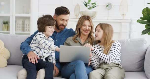 Cheerful small children sitting in hugs with parents on sofa and watching video on laptop computer. Mother father with little kids, daughter and son talking and applauding. Family on lockdown at home. — Stock Video