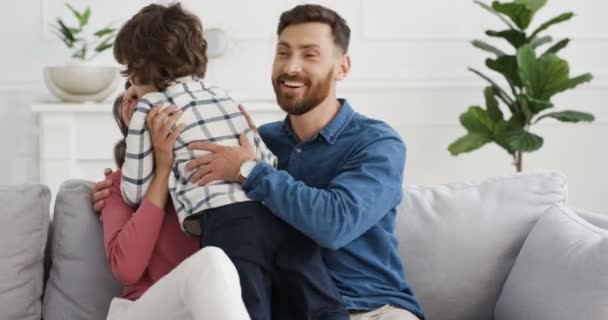 Caucasiano menino bonito correndo para a jovem mãe feliz e pai sentado no sofá na acolhedora sala de estar. Pais alegres abraçando e abraçando seu pequeno filho em casa no sofá. Interior . — Vídeo de Stock