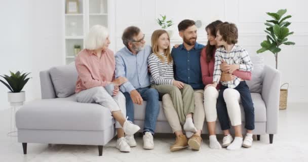 Portret białej szczęśliwej rodziny uśmiechniętej do kamery w przytulnym salonie. Radosny mężczyzna i kobieta siedzący na kanapie w uściskach z dziećmi i rodzicami. Dziadkowie, rodzice i dzieci w domu — Wideo stockowe