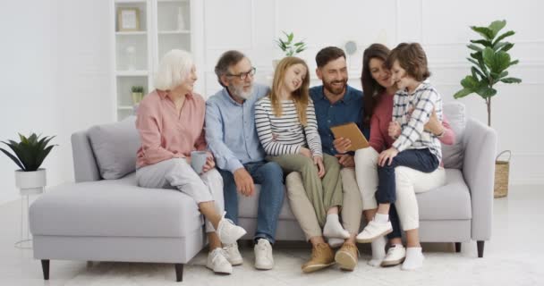 Caucasico felice grande famiglia sorridente e guardare video sul dispositivo tablet in camera accogliente. Gioioso uomo e donna seduti sul divano con bambini e genitori. Nonni, genitori e figli che utilizzano il computer. — Video Stock