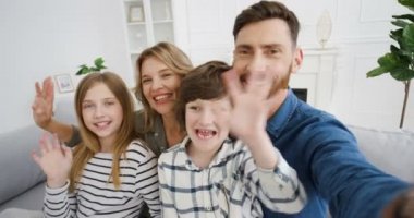 Kanepede oturan, gülümseyen ve kameraya el sallayan mutlu beyaz aile ve çocukların POV 'u. Anne ve babasıyla birlikte neşeli çocuklar web kameralarıyla konuşuyor ve evde görüntülü sohbet ediyorlar. Videochat çevrimiçi.