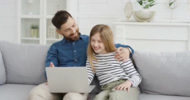 Genç beyaz bir baba oturma odasında kanepede oturuyor ve küçük şirin kızına dizüstü bilgisayar kullanmayı öğretiyor. Babasıyla bilgisayarda yazan küçük bir kız. Kapalı alanda. Baba ve çocuk evde oyun oynuyorlar..