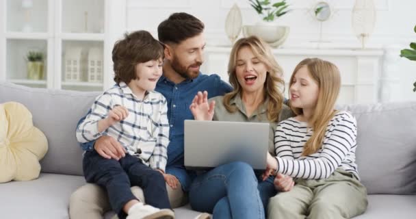 Szczęśliwa biała rodzina spędzająca wolny czas w domu. Małe słodkie dzieci z rodzicami rozmawiającymi i oglądającymi filmy na laptopie. Mama i ojciec dobrze się bawią z małym synem i córką przy komputerze. — Wideo stockowe