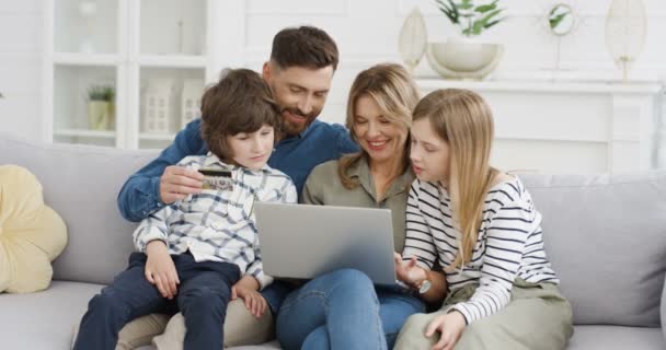 Biała rodzina z dziećmi wydającymi pieniądze na sofę w domu. Rodzice z dziećmi korzystającymi z laptopa do zakupu online za pomocą karty kredytowej. Dzieci z matką i ojcem zakupy w Internecie na komputerze. — Wideo stockowe