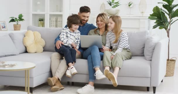 Los padres caucásicos con niños en el sofá usando gadget. Joven hombre y mujer con hijo e hija pasar tiempo junto con el ordenador portátil. Padre y madre viendo algo en línea en la computadora. Descanse en casa . — Vídeos de Stock