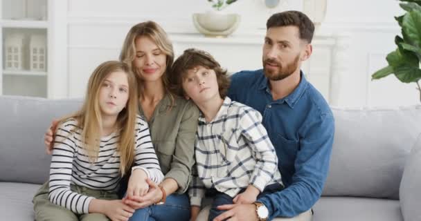 Portrait of cheeful Caucasian family with two kids sitting on couch and smiling joyfully to camera. Small cute boy and girl with mother and father at home. Parents with little children hugging. — Stock Video