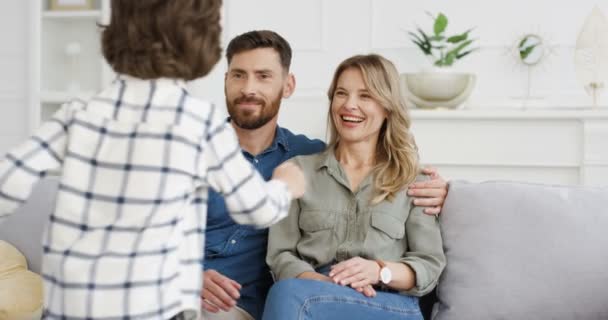 Rear of Caucasian cute little kid boy running to young happy mother and father sitting on couch in cozy living room. Cheerful parents hugging and embracing their small son at home on sofa. Back view. — Stock Video