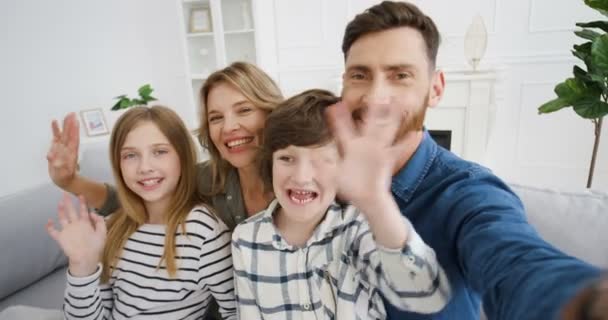 POV di genitori caucasici felici e bambini seduti sul divano, sorridenti e salutando allegramente alla macchina fotografica. Bambini gioiosi con madre e padre che parlano con webcam e videochat a casa. Videochat online. — Video Stock