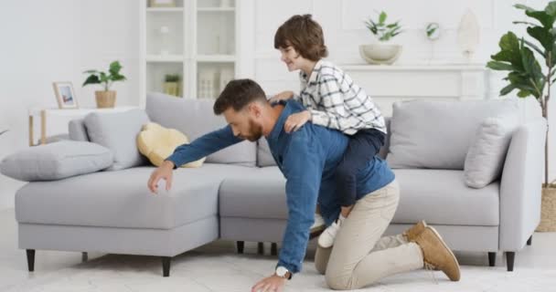 Alegre padre caucásico feliz jugando con pequeño hijo lindo en la sala de estar. Papá sosteniendo a un niño pequeño en la espalda y arrastrándose por el suelo en casa. Cabalgando sobre papá de vuelta. Padres divirtiéndose con el niño . — Vídeos de Stock