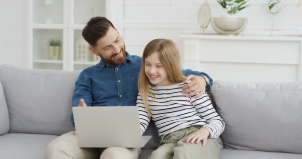 Młody biały ojciec siedzi na kanapie w salonie i uczy małą, słodką córkę za pomocą laptopa. Mała dziewczynka pisząca na komputerze ze swoim ojcem. W środku. Tatuś i dziecko bawią się online w domu. — Wideo stockowe
