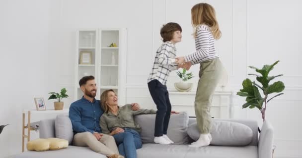 Cute small Caucasian boy and girl playing and jumping on couch in living room. Parents smiling and sitting on sofa beside. Children having fun at home. Mother and father watching their kids. — Stock Video