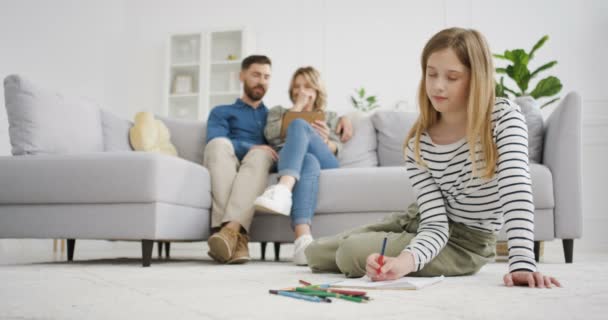 Kafkasyalı mutlu çocuk yerde oturuyor ve renkli kalemlerle resim boyuyor. Evde oynayan ve resim çizen küçük tatlı bir kız. Ebeveynler kanepeye oturur ve arka planda konuşurlar. Anne ve baba dinleniyor. — Stok video