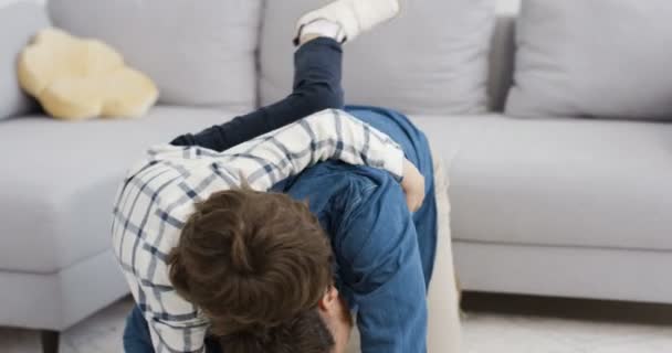 Feliz hombre caucásico alegre abrazando a su pequeño hijo lindo y riendo en casa en el suelo. Padres alegres jugando y divirtiéndose con niños pequeños en la sala de estar. Joven padre luchando alegremente con el niño . — Vídeos de Stock