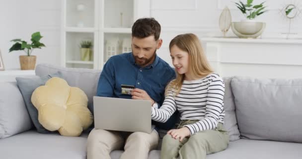 白人父亲和可爱的女儿坐在沙发上，用笔记本电脑，用信用卡在网上购物。小女孩和她爸爸在网上买东西。室内。孩子和爸爸一起花钱. — 图库视频影像