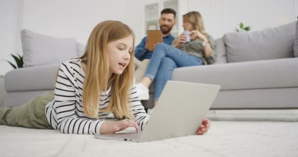 Kaukasisch gelukkig kind liggend op de vloer en typend op toetsenbord van laptop. Klein schattig meisje dat thuis op de computer speelt. Ouders zitten op de bank en praten op de achtergrond. Moeder en vader rusten uit. — Stockvideo