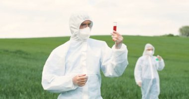 Beyaz koruyucu takım elbiseli, gözlüklü, yeşil alanda kırmızı kimyasal sıvıyla test tüpü tutan ve böcek ilaçlarını araştıran Kafkas bilim adamı. Arka planda kadın biyolog.