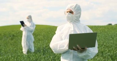 Beyaz koruyucu kostümlü, gözlüklü, yeşil alanda duran ve dizüstü bilgisayar üzerinde çalışan beyaz erkek bir çiftçi biyoloğu. İnsan tarım ekolojisti ve bilim adamı hasatı araştırıyor.