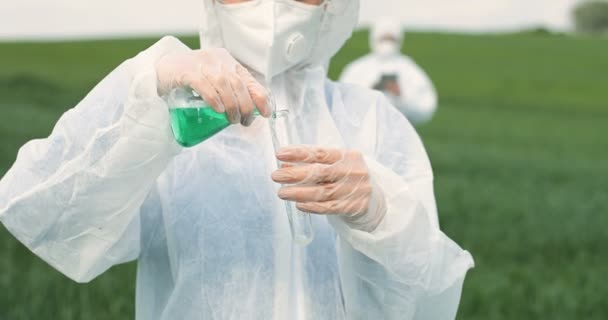 Primo piano del tubo del tet con liquido chimico nelle mani di ricercatrice caucasica femminile che si trova in campo verde. pesticidi agricoli che utilizzano. Donna in tuta protettiva che fa controllo dei parassiti. — Video Stock