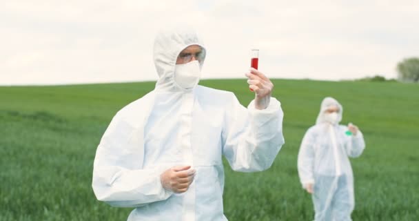 Caucasian man scientist in white protective suit and goggles holding test tube with red chemical liquid in green field and exploring pesticides. Woman biologist on background. — Stock Video