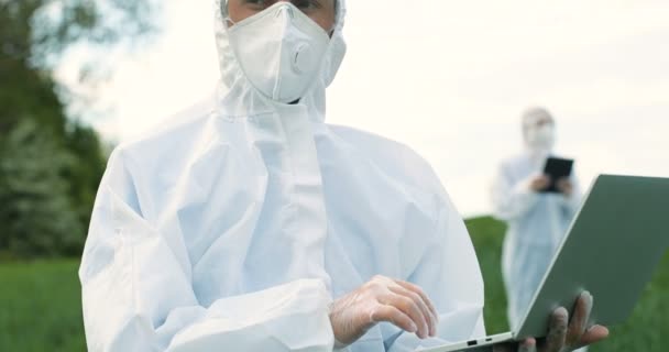 白い保護衣装、マスクやゴーグル緑のフィールドに立って、ラップトップコンピュータ上で動作する男性白人農家の生物学者。人間の農業生態学者収穫と害虫を探る科学者. — ストック動画