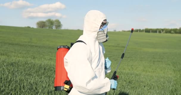 Portrait of Caucasian male farmer in white protective clothes, gloves, mask and goggles standing in green field with pulverizator for spraying pesticides. Man with chemicals for fumigation of harvest. — Stock Video