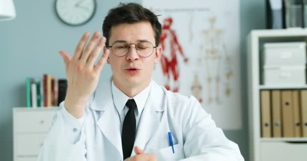 Caucasiano jovem bonito médico em vestido branco e óculos sentados à mesa no armário e explicando algo para a câmera. Homem blogueiro médico falando sobre cuidados de saúde online. Videoblog médico . — Vídeo de Stock