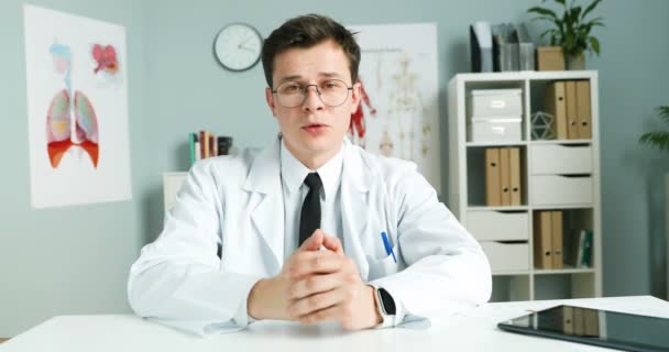 Mężczyzna lekarz blogger w białej sukni i okulary siedzi przy biurku w gabinecie i opowiada o opiece zdrowotnej online. Bloguję o zdrowiu. Lekarz prowadzący konsultacje w Internecie. Konsultacje medyczne w vlog. — Wideo stockowe
