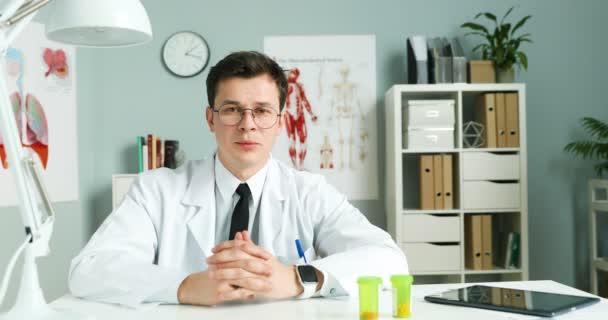 Médico jovem caucasiano de vestido branco e óculos sentados na mesa no escritório, mostrando pílulas e remédio para a câmera. Homem blogueiro médico falando sobre saúde e medicina online. Videoblog médico . — Vídeo de Stock
