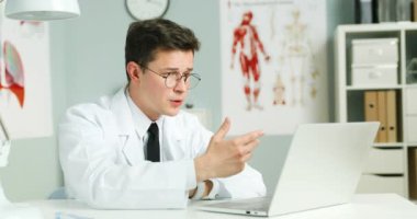 Beyaz önlüklü ve gözlüklü beyaz erkek doktor dolapta masada oturuyor. Doktor dizüstü bilgisayarda görüntülü konuşma yapıyor ve hastaya webcam üzerinden danışmanlık yapıyor. Medic video kaset ve danışmayı çevrimiçi yapıyor.