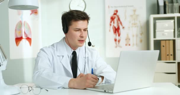 Άνδρας όμορφος Καυκάσιος γιατρός με ακουστικά κάθεται στο τραπέζι στο ντουλάπι. Γιατρός videochatting σε φορητό υπολογιστή και τη διαβούλευση ασθενή μέσω κάμερας. Γιατρός που έχει βιντεοκλήση και διαβούλευση στο διαδίκτυο. — Αρχείο Βίντεο