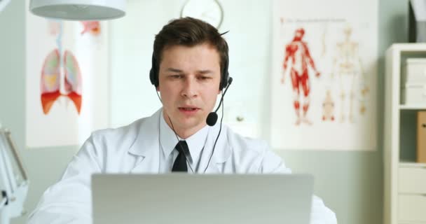 Άνδρας όμορφος Καυκάσιος γιατρός με ακουστικά που κάθεται στο γραφείο του. Γιατρός videochatting σε φορητό υπολογιστή και τη διαβούλευση ασθενή μέσω κάμερας. Γιατρός που έχει βιντεοκλήση και διαβούλευση στο διαδίκτυο. — Αρχείο Βίντεο